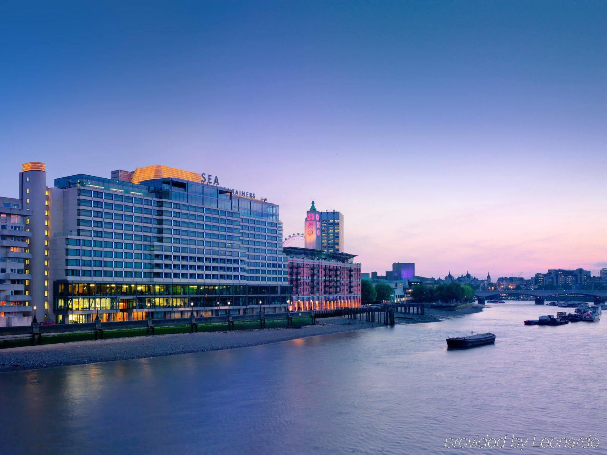 מלון Sea Containers לונדון מראה חיצוני תמונה The photo depicts a waterfront scene featuring a modern hotel building next to a river. The hotel has a sleek, contemporary design, with multiple stories and large windows reflecting the evening light. The sky transitions from a soft blue to warmer h
